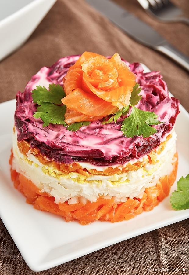 Salad cake with layers of carrots, beets, boiled egg and smoked salmon on a white plate.