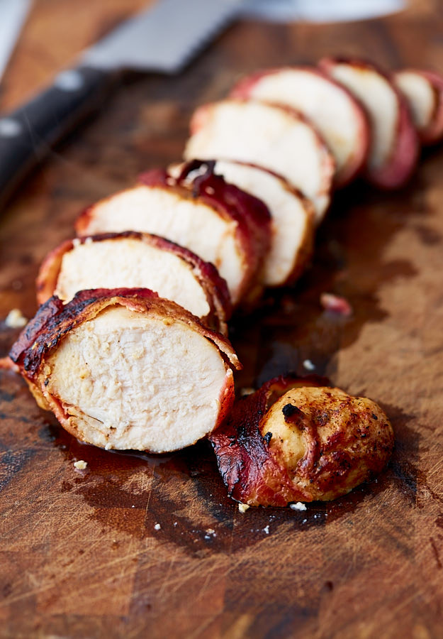 Sliced crispy bacon-wrapped chicken breast on a brown cutting board.