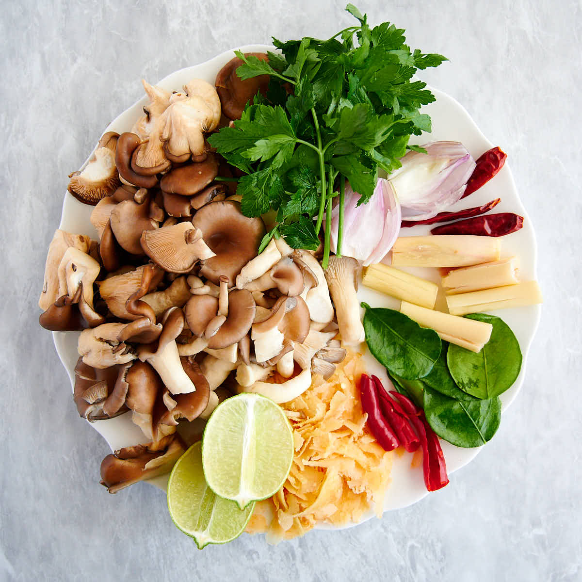 Mushrooms, galangal, lime, lime leaves, shallots, lemongrass, Thai chiles and cilantro on a white plate.